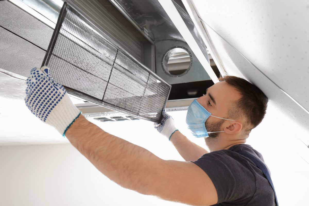 A man in a mask and gloves repairs an air conditioner, emphasizing air duct cleaning for improved indoor air quality.